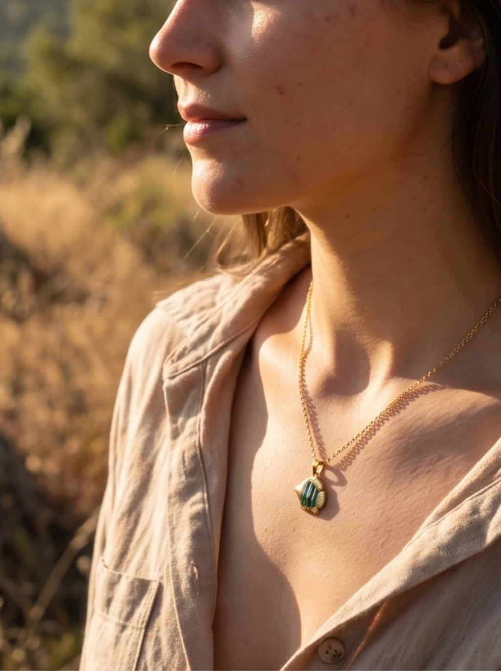 Side profile lifestyle image of a model wearing the fish pendant necklace against a soft-focus outdoor field at golden hour, exuding organic coastal warmth