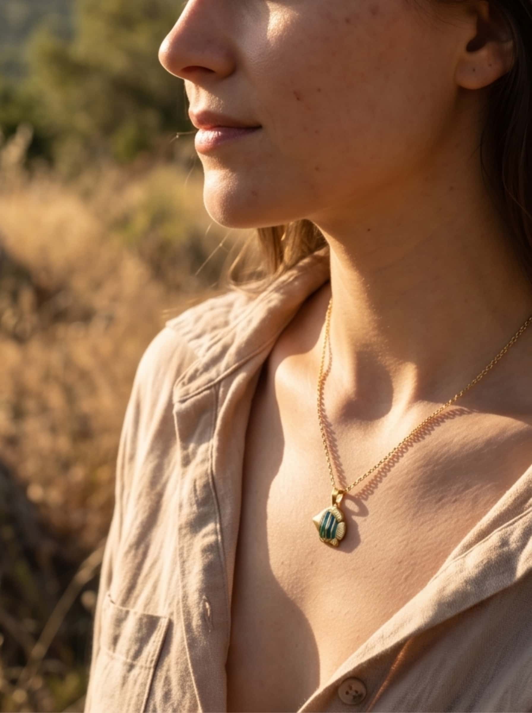 Side profile lifestyle image of a model wearing the fish pendant necklace against a soft-focus outdoor field at golden hour, exuding organic coastal warmth