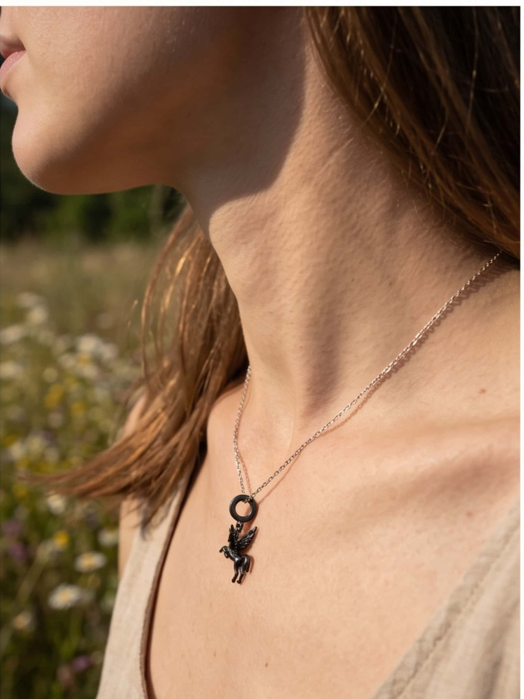Side profile of a woman standing in a sunlit wildflower field wearing the dark sterling silver unicorn necklace, showcasing how the pendant drapes naturally