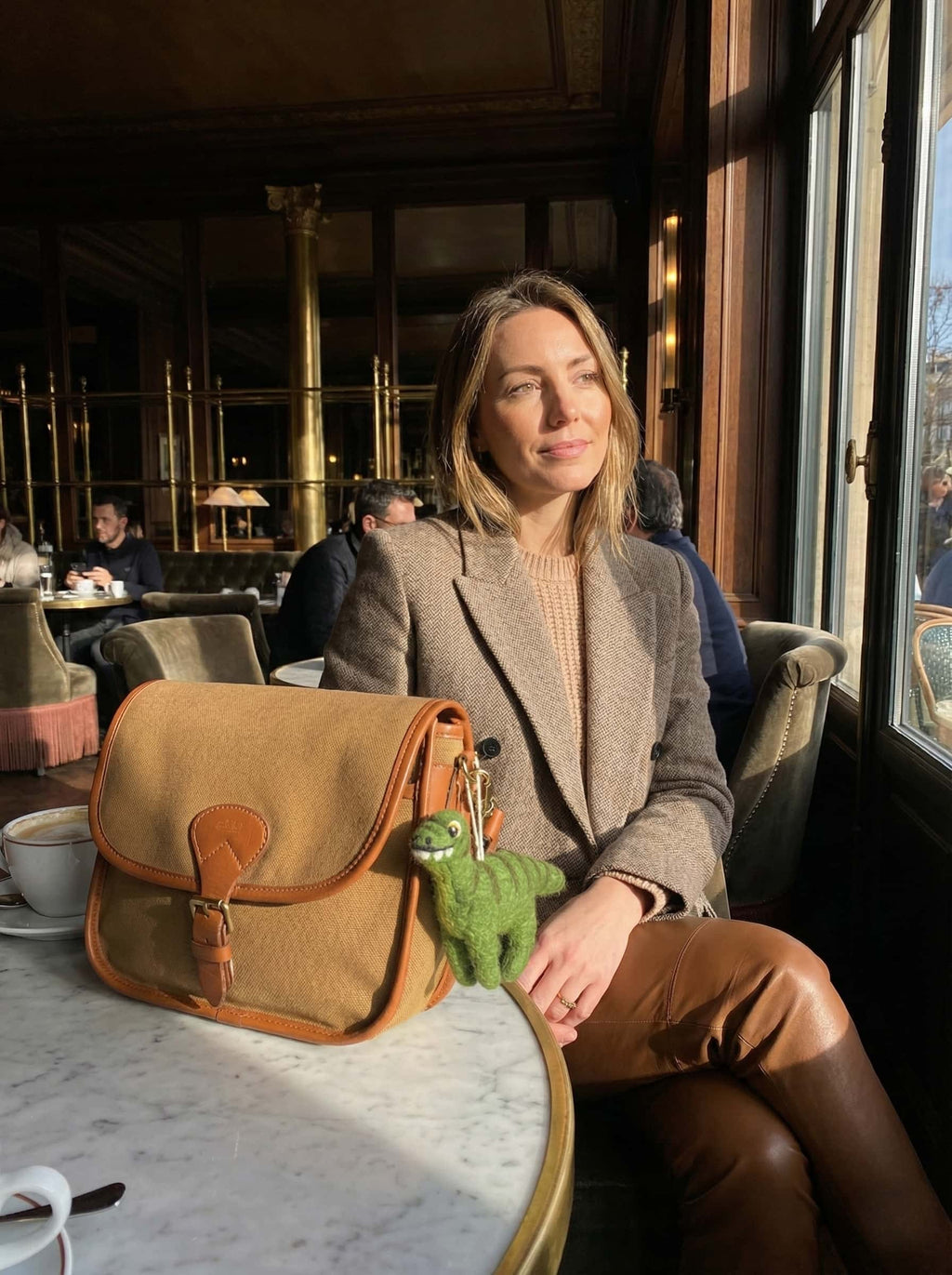 Woman sitting at a cafe table with a brown leather bag and green wool felt ornament dinosaur on a marble surface.