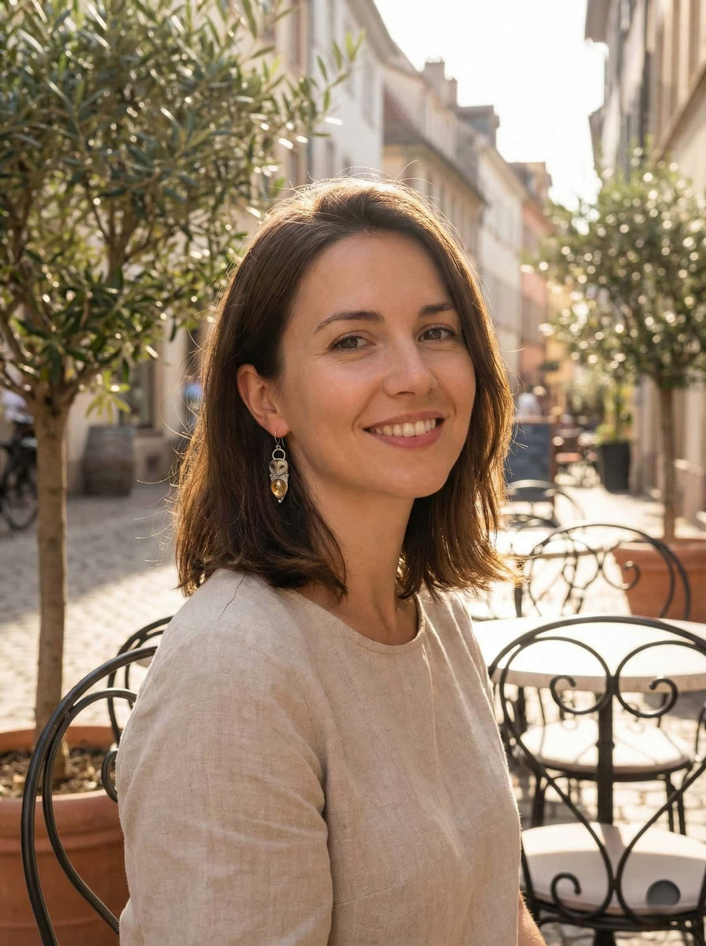 Woman wearing owl earrings sitting outdoors in a sunlit street with trees and buildings in the background