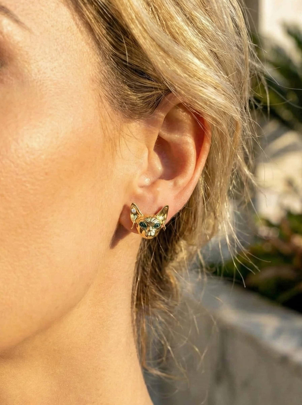 Close-up of a person wearing a gold sphinx-cat earrings outdoors.