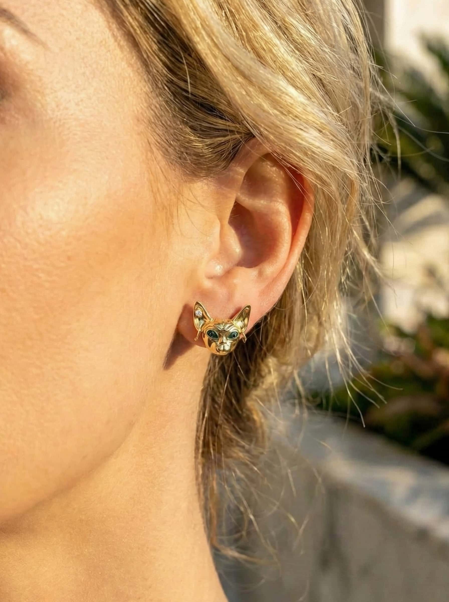 Close-up of a person wearing a gold sphinx-cat earrings outdoors.
