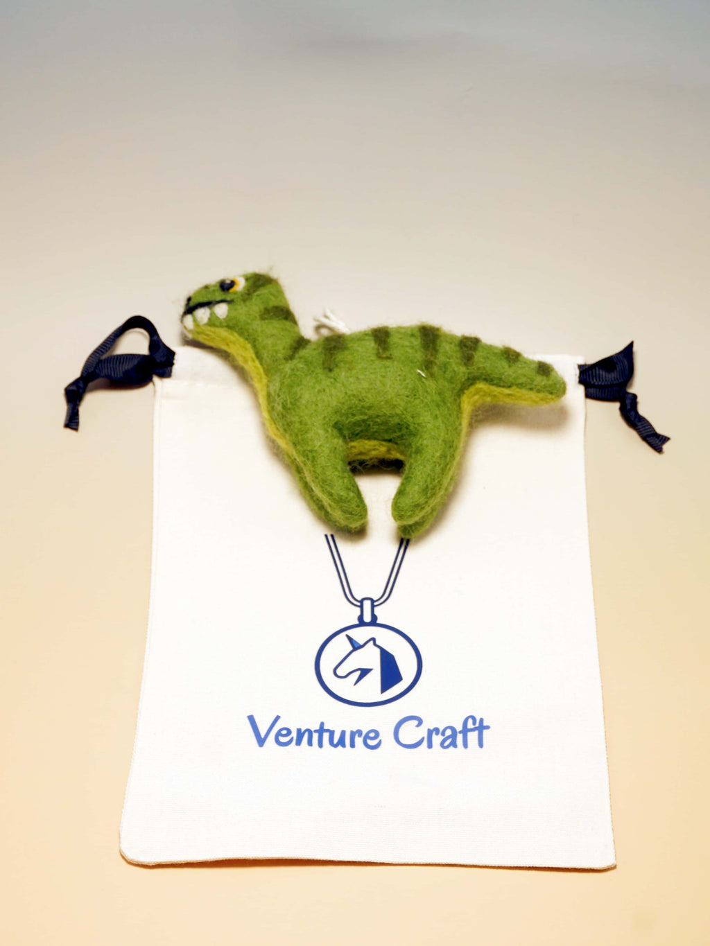 Green needle felted dinosaur with a 'Venture Craft' logo on a beige background