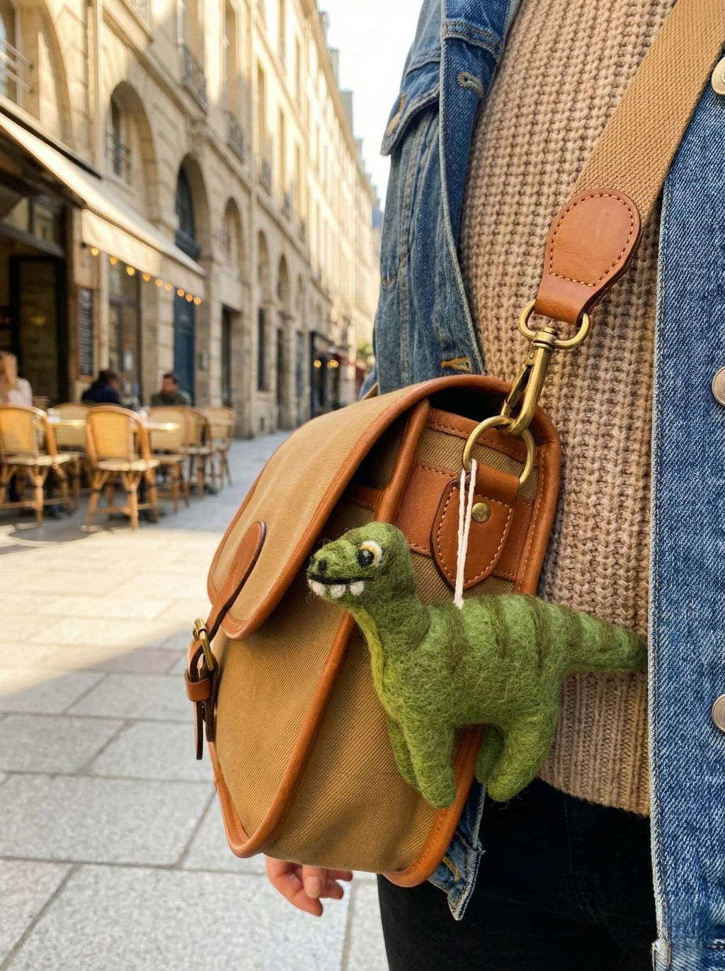 Person holding a brown leather bag with a green wool felt ornament dinosaur on a city street.