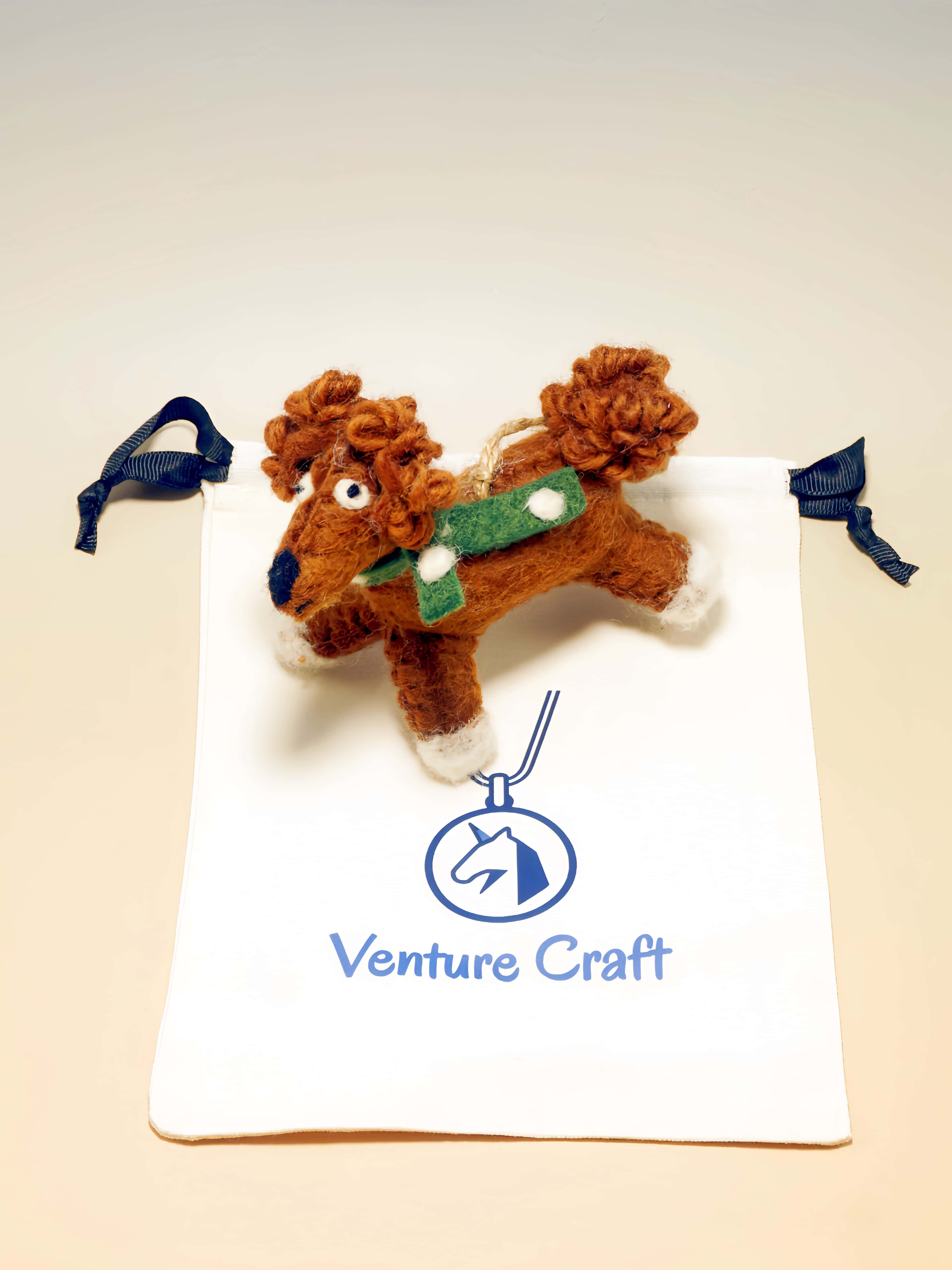 Brown wool felt dog ornament with a green saddle blanket on a white pillow, branded 'Venture Craft'.