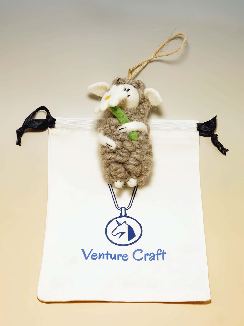 Plush wool felt sheep ornament with flower hanging from a string on a Venture Craft branded pouch.