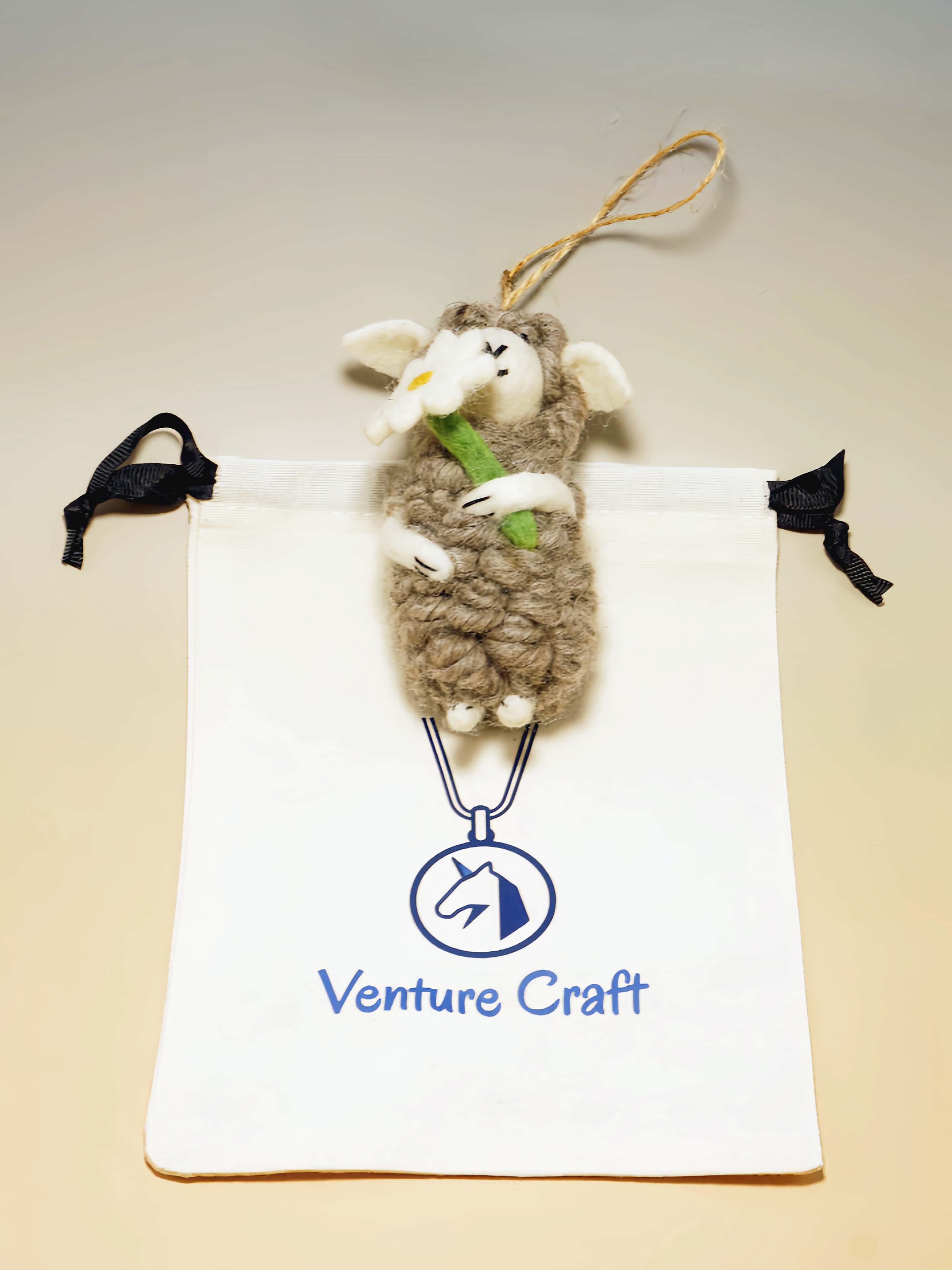 Plush wool felt sheep ornament with flower hanging from a string on a Venture Craft branded pouch.