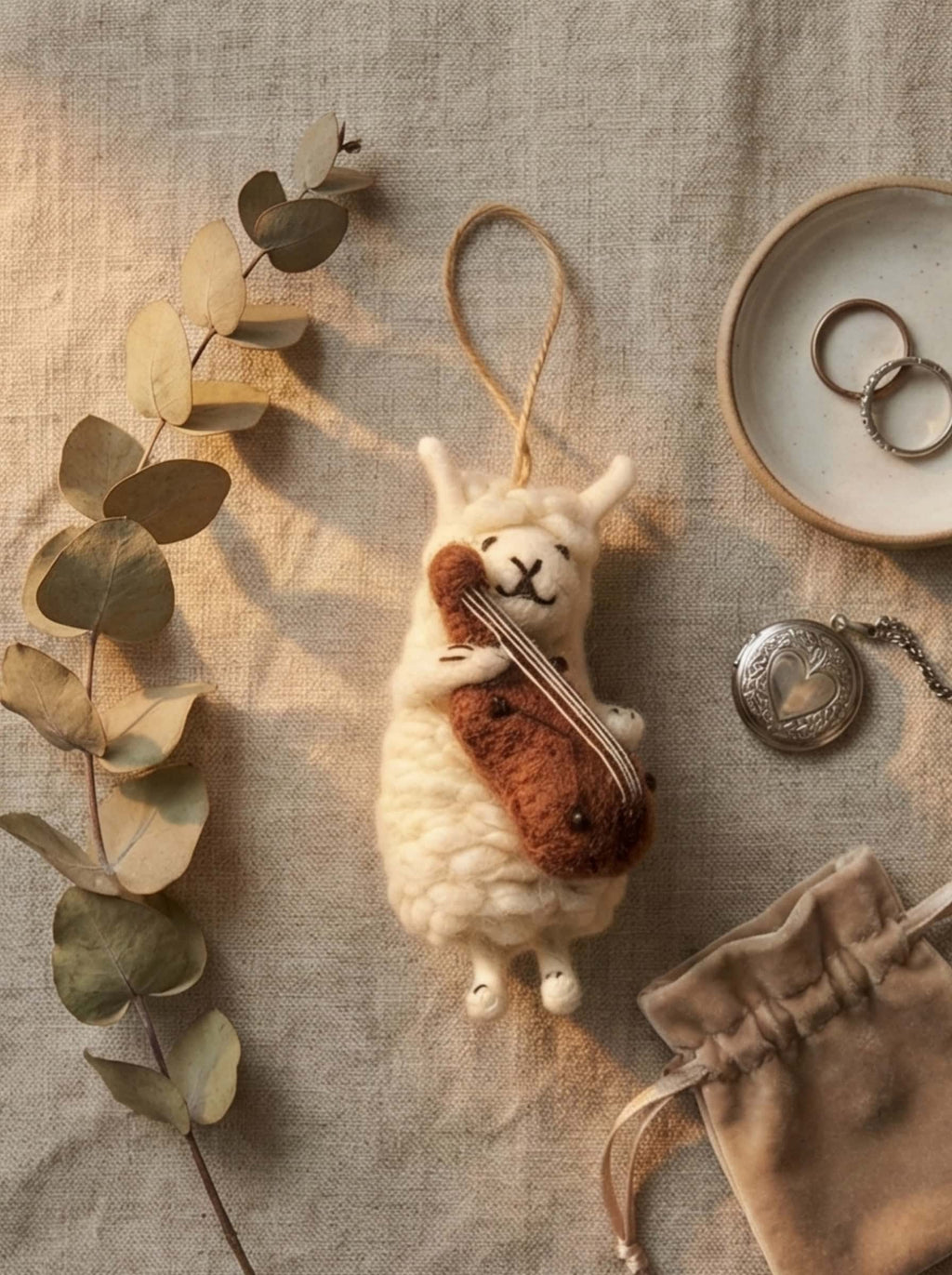 Small wool felt sheep with violin on a textured surface with eucalyptus leaves and jewelry.