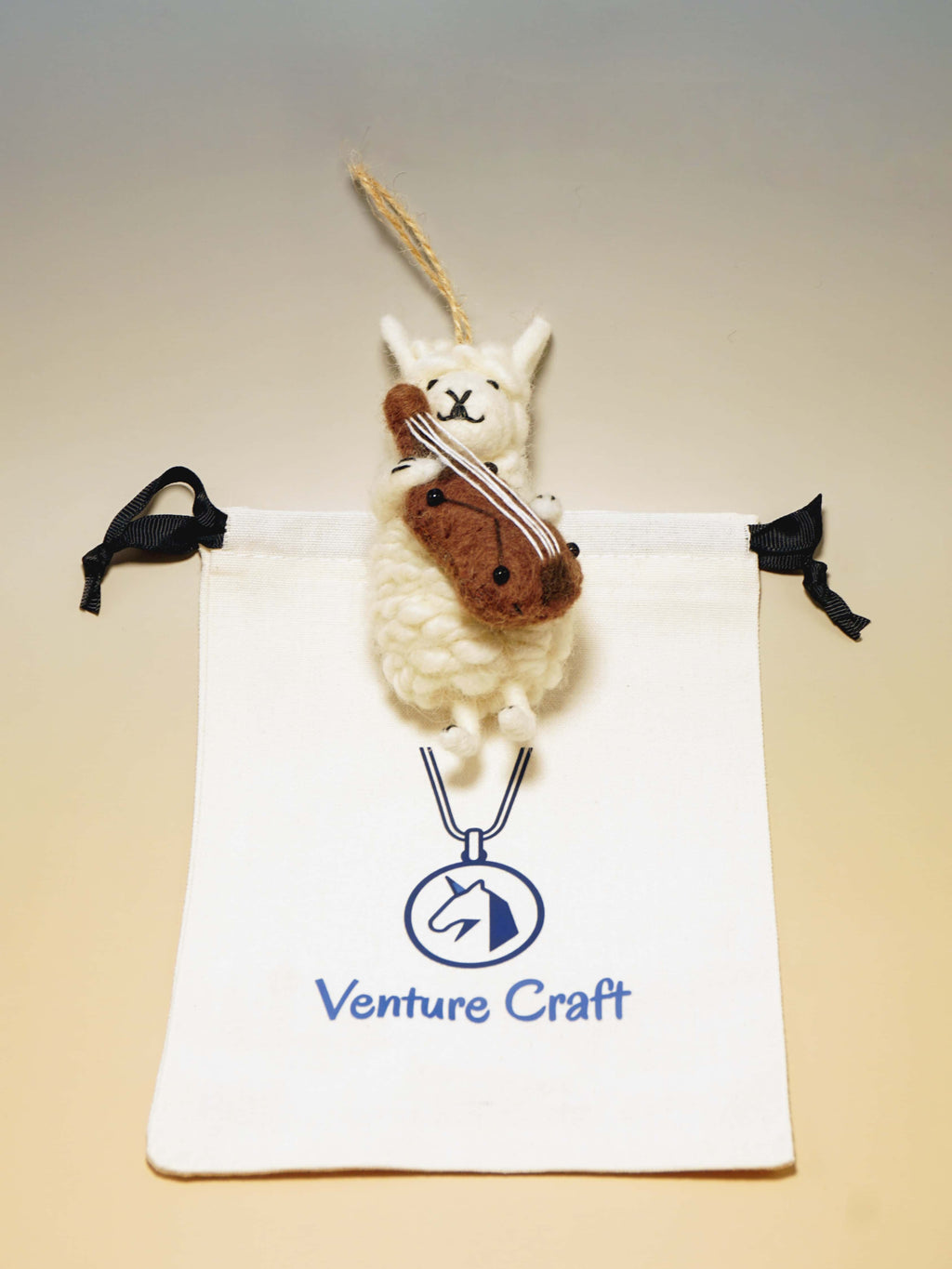 Small wool felt sheep with violin hanging from a string on a Venture Craft branded pouch.