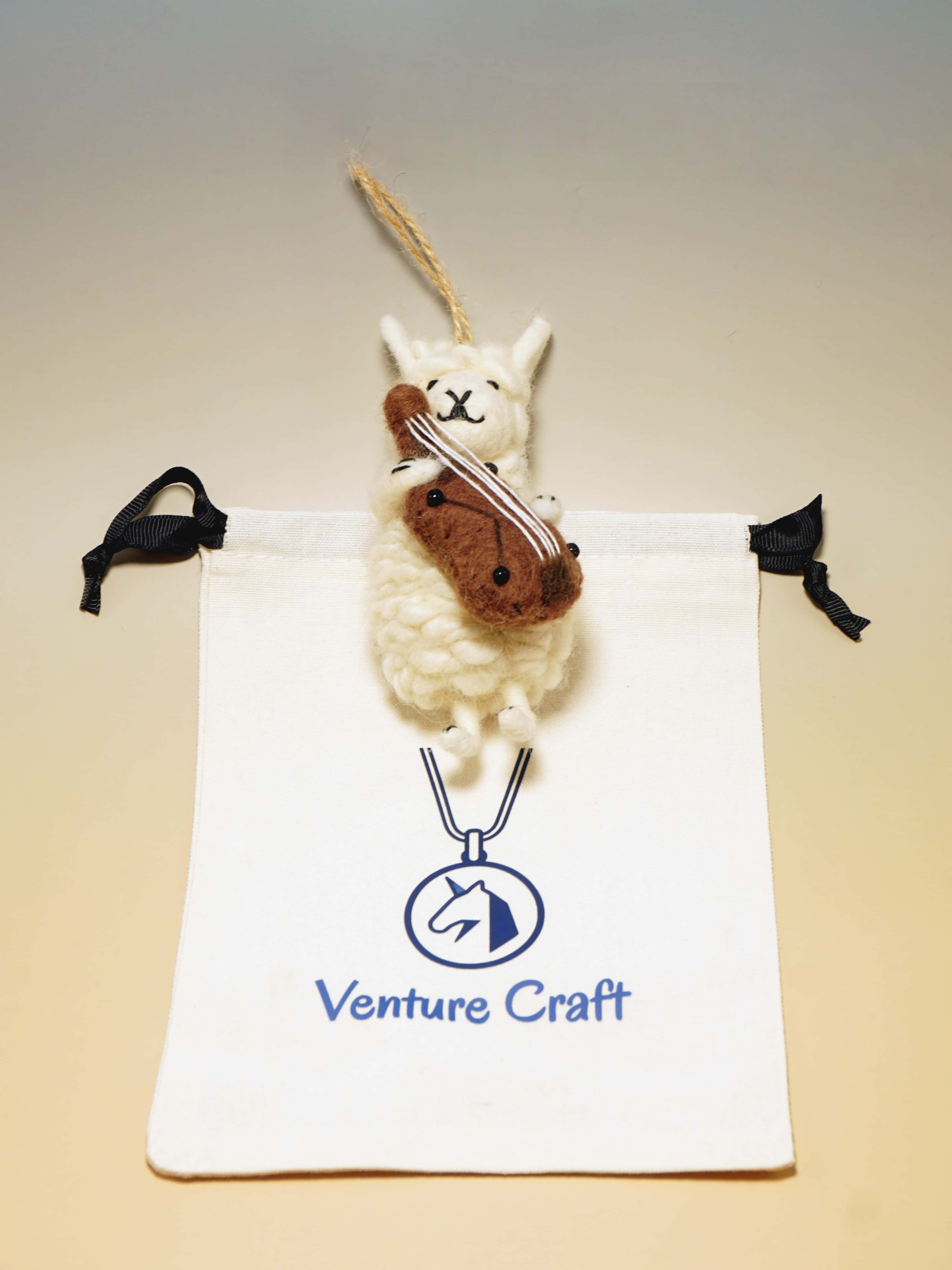 Small wool felt sheep with violin hanging from a string on a Venture Craft branded pouch.
