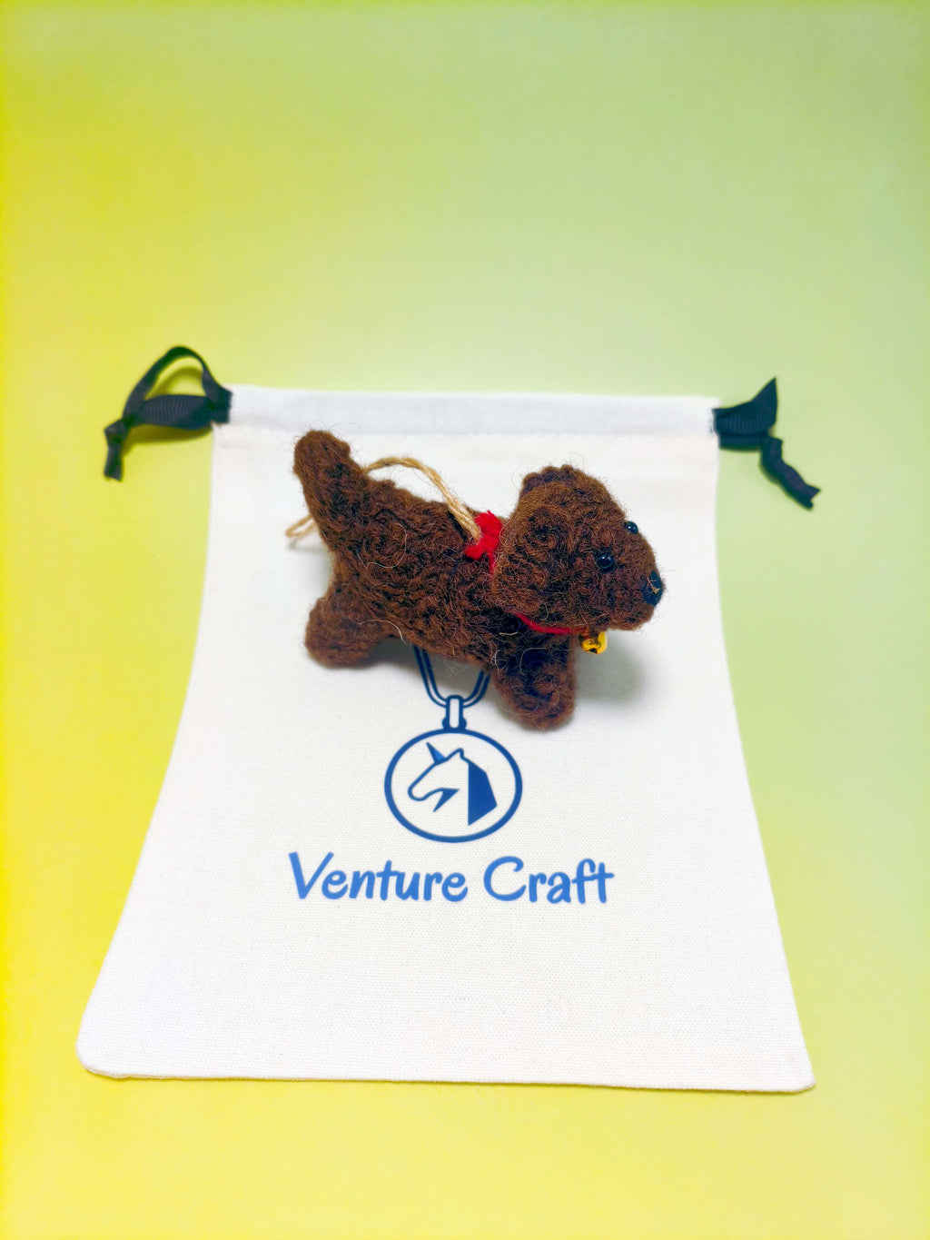 Crocheted wool felt toy poodle ornament with a red bow on a white drawstring bag with 'Venture Craft' logo against a yellow gradient background.